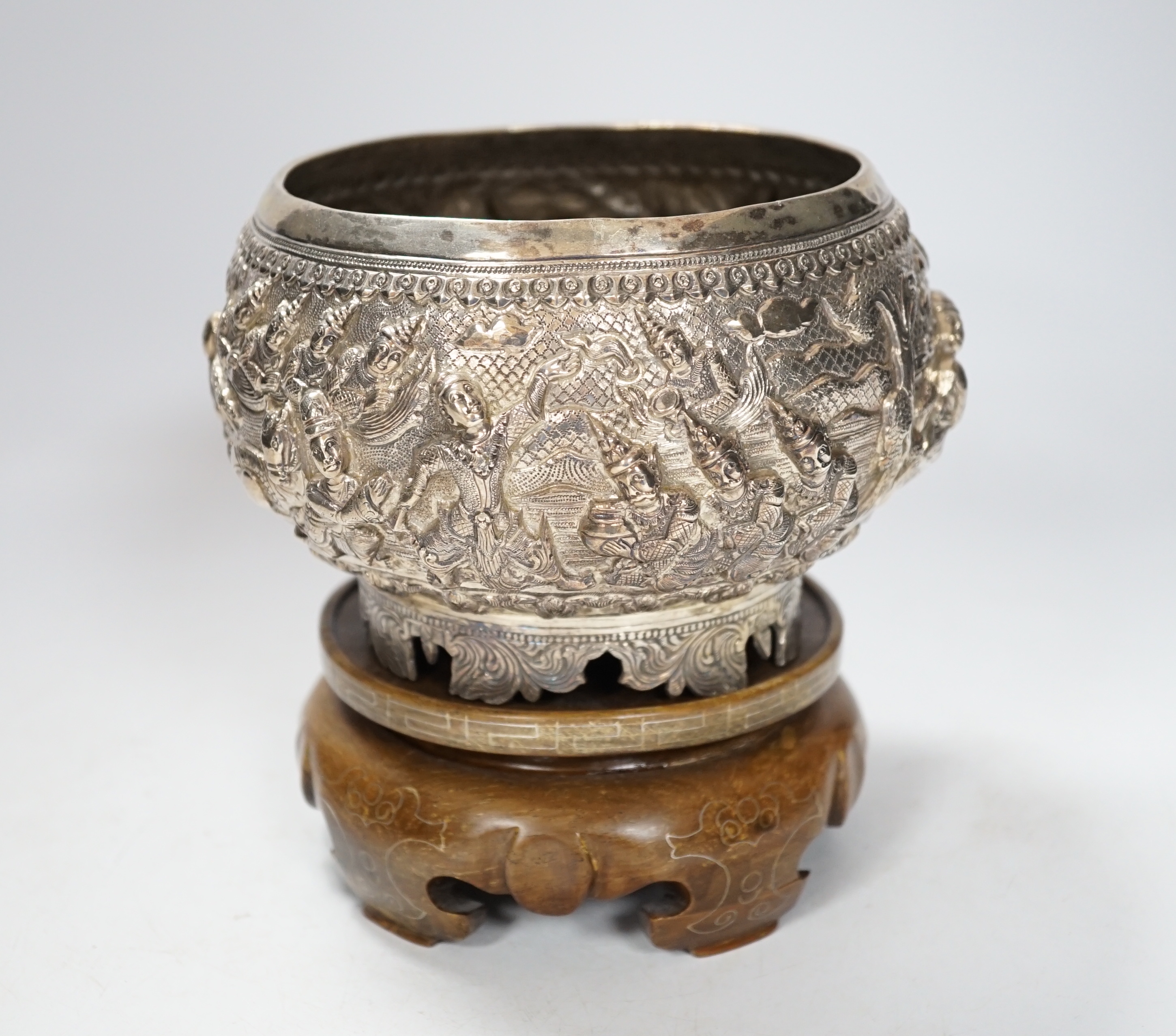 An Indian circular white metal bowl, decorated with a continuous band of figures in a landscape, on scrolled feet with hardwood stand, diameter 16cm